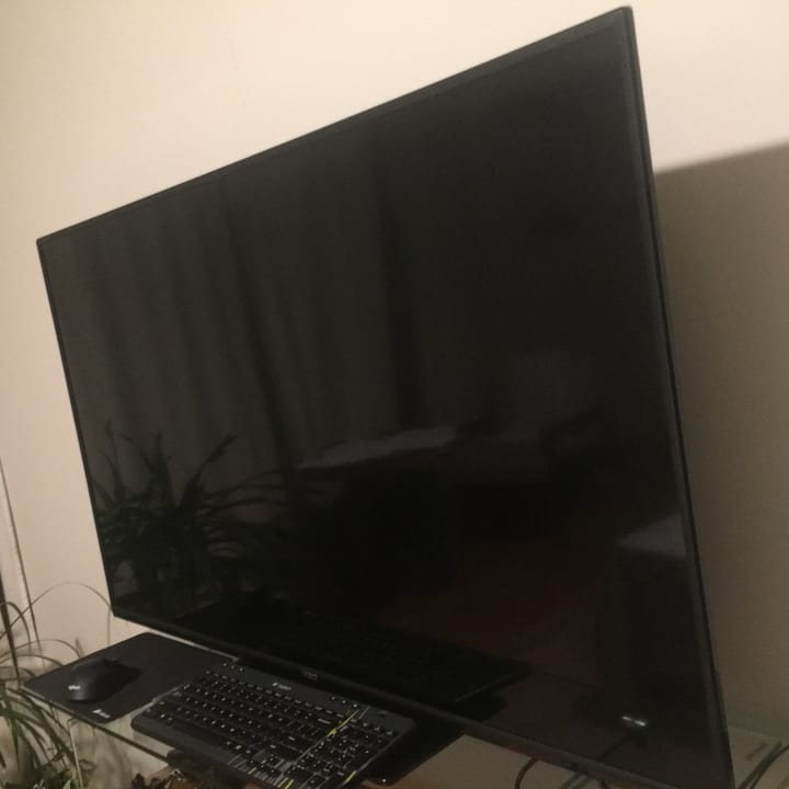 Photo of large, all black, wide-screen TV, with black keyboard, mouse, and mousepad on narrow glass table against white wall