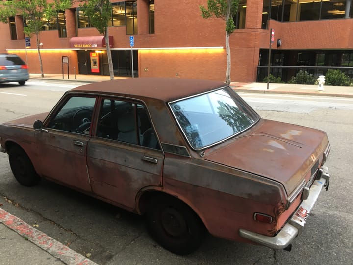 Photo of rusted orange car in foreground with orange bbank building in background